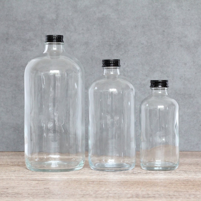 May include: Three clear glass bottles with black lids. The bottles are different sizes and are arranged in a row from largest to smallest.