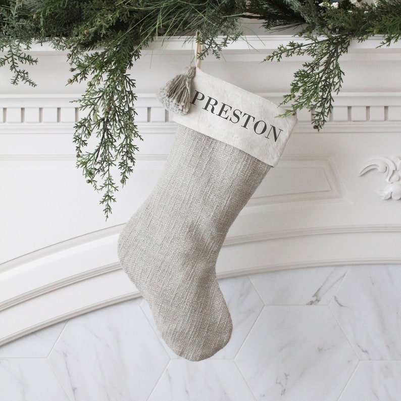 May include: A gray Christmas stocking with the name "PRESTON" printed on the top. The stocking is hanging from a white mantelpiece decorated with greenery. The stocking has a textured pattern and a gray tassel.