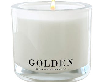 Puede incluir: Una vela blanca en un tarro de vidrio transparente con una etiqueta negra que dice "GOLDEN MANGO + DRIFTWOOD".