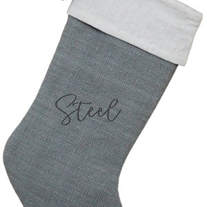 May include: Gray Christmas stocking with a white cuff and a gray tassel. The word "Steel" is written in cursive on the front of the stocking. The stocking has a loop for hanging.