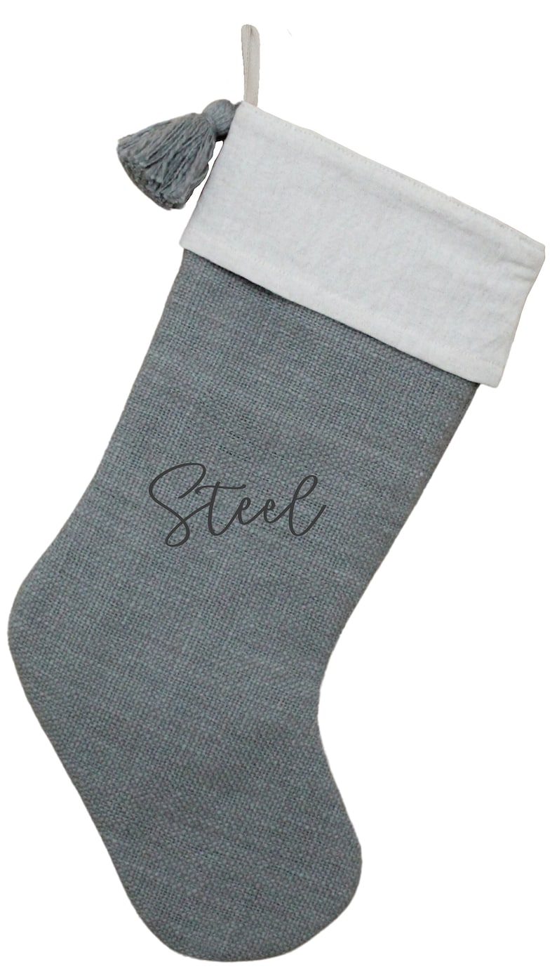 May include: Gray Christmas stocking with a white cuff and a gray tassel. The word "Steel" is written in cursive on the front of the stocking. The stocking has a loop for hanging.