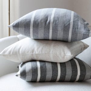 May include: Four square cushions stacked on top of each other. Each cushion features a striped design with alternating wide and thin vertical stripes. The colour palette is grey and white, offering a neutral and contemporary look.