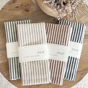 May include: Four folded striped tea towels in shades of gray, beige, and brown are displayed. Each towel is wrapped with a white paper band that reads "PLH STRIPE TEA TOWEL". The towels are made of premium cotton and linen blend.
