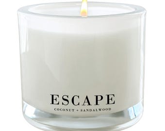 Puede incluir: Una vela blanca en un tarro de vidrio transparente con una etiqueta negra que dice "ESCAPE COCONUT + SANDALWOOD". La vela está encendida y la llama es visible.
