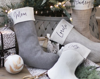 May include: Four personalised Christmas stockings made from grey and white fabric. The stockings are decorated with tassels and have the names "Mom", "Audrey", "Lorelei", and "Lily" written on them.