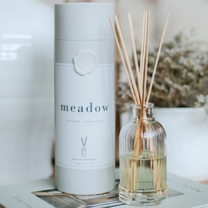 Puede incluir: Un juego de difusor de aceite de caña con una botella de vidrio transparente y un recipiente cilíndrico. La botella contiene un líquido de color claro y cañas de madera. El recipiente es blanco con la palabra "meadow" y la descripción de la fragancia "Juniper + Bergamot".
