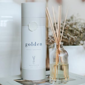 May include: A white reed oil diffuser set with the word "golden" printed on the label. The set includes a glass bottle with reeds and a cylindrical container. The label also reads "Mango + Driftwood" and "Reed Oil Diffuser 6 oz / 177 ml."