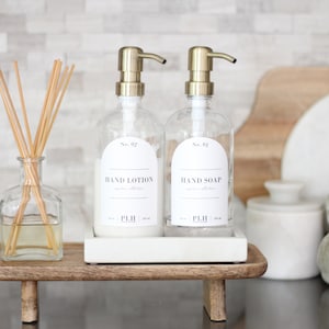 May include: Two clear glass pump bottles with gold tops and white labels on a wooden tray. The labels read "No. 01 HAND SOAP" and "No. 02 HAND LOTION".