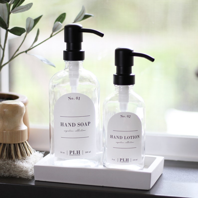 Op de afbeelding: Twee doorzichtige glazen zeepdispensers met zwarte pompen, met de labels "Hand Soap" en "Hand Lotion". De dispensers staan op een wit dienblad. De handzeepdispenser is 500 ml, en de handlotiondispenser is 250 ml. De labels zijn wit met zwarte tekst.