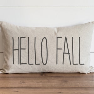 Op de afbeelding: Een beige kussen met de tekst "HELLO FALL" in zwarte letters.