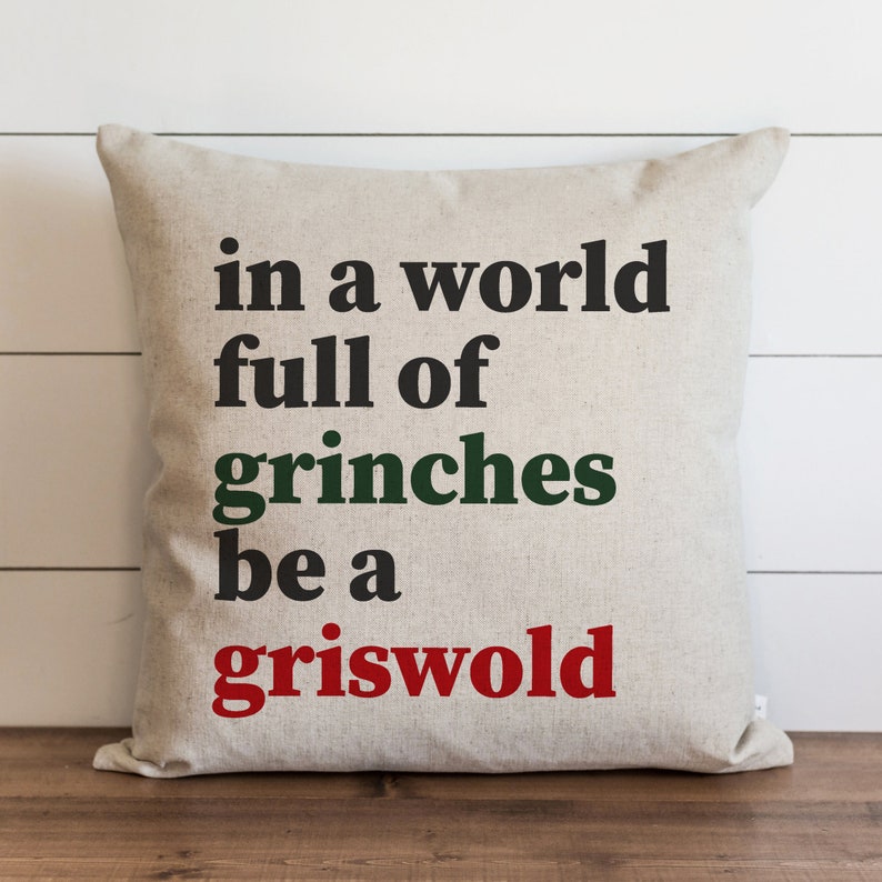 Puede incluir: Un coj&iacute;n cuadrado beige con la frase "in a world full of grinches be a griswold" en negro, verde y rojo. El coj&iacute;n est&aacute; hecho de una tela texturizada, adecuado para la decoraci&oacute;n del hogar.