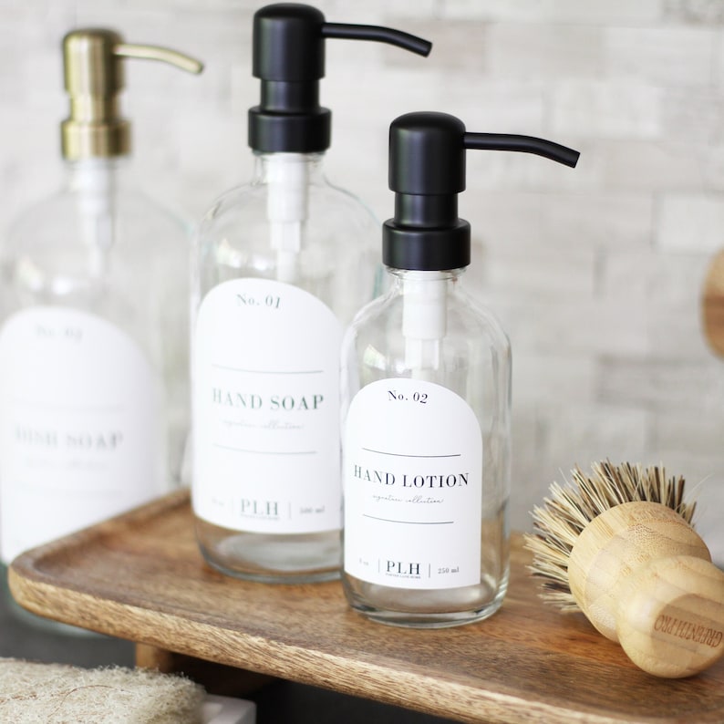 May include: Three glass bottles with black pump tops and white labels. The labels have black text that reads "No. 01 Hand Soap", "No. 02 Hand Lotion", and "No. 03 Body Soap". The bottles are sitting on a wooden tray with a wooden brush.