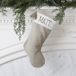 May include: A beige Christmas stocking with the name "MATT" printed in black on a white cuff. The stocking is made of a textured fabric and hangs from a decorative garland. A tassel hangs from the top of the stocking.