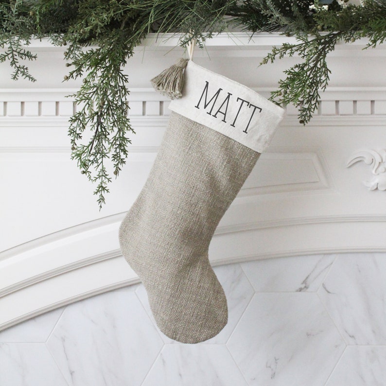 May include: A beige Christmas stocking with the name "MATT" printed in black on a white cuff. The stocking is made of a textured fabric and hangs from a decorative garland. A tassel hangs from the top of the stocking.