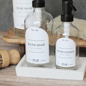 Op de afbeelding: Transparante glazen zeep- en lotiondispensers met zwarte pompen en witte etiketten. De etiketten luiden "Handzeep" en "Handlotion". De zeepfles is 500 ml, en de lotionfles is 250 ml. De flessen staan op een wit dienblad.