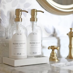 May include: Two gold pump bottles with clear glass containers and white labels. The labels read "No. 01 HAND SOAP" and "No. 02 HAND LOTION". The bottles are on a white rectangular tray.