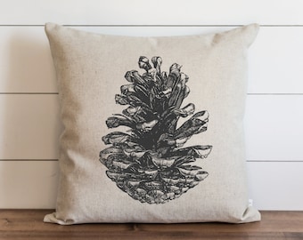 May include: A beige pillow with a black and white illustration of a pine cone.