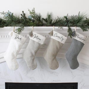 May include: Four Christmas stockings in varying shades of white, gray, and beige hang from a mantel. Each stocking is personalized with a name: "Dad," "Mom," "Everly," and "Preston." The stockings are adorned with tassels and are decorated with greenery.