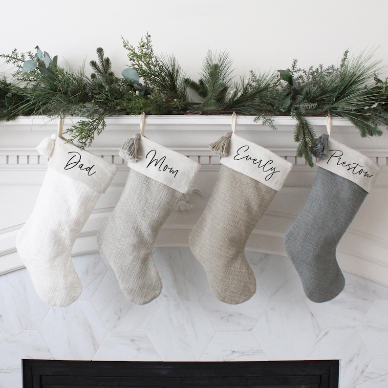 May include: Four Christmas stockings in varying shades of white, gray, and beige hang from a mantel. Each stocking is personalized with a name: "Dad," "Mom," "Everly," and "Preston." The stockings are adorned with tassels and are decorated with greenery.