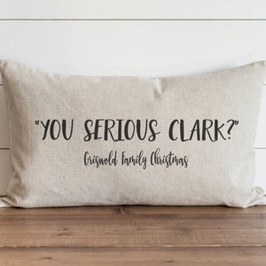 Op de afbeelding: Een beige kussen met de tekst "YOU SERIOUS CLARK?" en "Griswold Family Christmas" in zwarte letters.