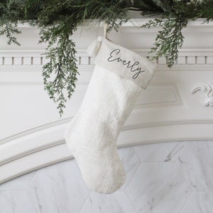 May include: A cream-colored Christmas stocking with the name "Everly" in black script. The stocking is hanging from a white mantelpiece, adorned with greenery. The stocking has a tassel and a textured fabric.