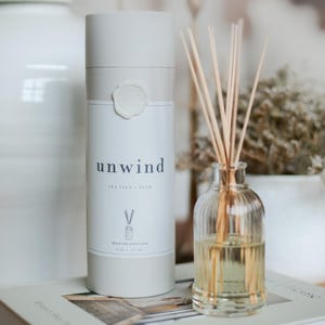 May include: A reed oil diffuser with the word "unwind" printed on the label. The diffuser includes a glass bottle with wooden reeds and a cylindrical container. The label reads "Sea Salt - Palo" and "Reed Oil Diffuser 177 ml."