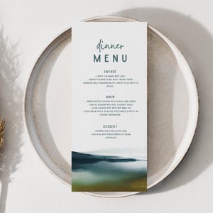 May include: A dinner menu card with a watercolor landscape design, featuring a white background and a teal and gold color scheme. The menu is placed on a white plate with a beige rim, and a dried plant is on the left.