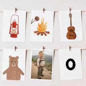 Op de afbeelding: Zes rechthoekige kaarten hangend aan een touwtje. De kaarten bevatten illustraties van een rode lantaarn, een kampvuur met een marshmallow, een bruine akoestische gitaar, een bruine beer, een foto van een kind en de zwarte letter O.