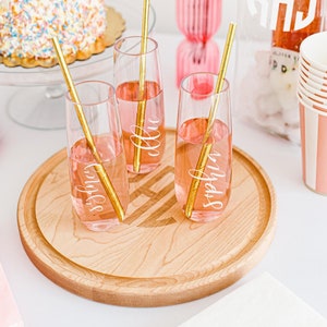 Könnte beinhalten: Drei Champagnerflöten mit personalisierten Namen "Sophia", "Ellie" und "Sydney" auf einem Holztablett. Die Flöten sind mit einem rosafarbenen Getränk gefüllt und haben goldene Strohhalme. Im Hintergrund befinden sich ein rosa-weiß gestreiftes Papierbecher und eine Flasche Champagner.