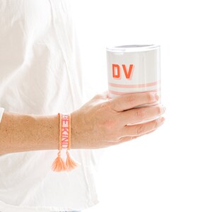 May include: A white and pink tumbler with the letters "DV" in red is held in the hand. A woven bracelet with the words "BE KIND" in orange is worn on the wrist. The bracelet has orange tassels. The person is wearing a white shirt and blue jeans.