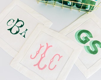 May include: A collection of white linen monogrammed coasters with embroidered initials in green and pink. A gold wire basket holds several green glass bottles of sparkling water, each with a blue label. Two clear drinking glasses are also visible.