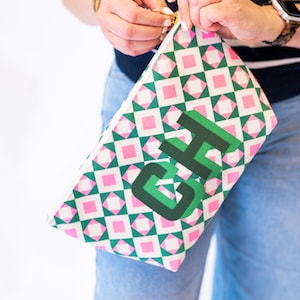 May include: A rectangular clutch with a geometric pattern of pink and green diamonds on a white background. The letters "GH" are printed in green on the front. The clutch has a gold zipper.