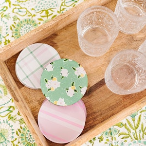 Könnte beinhalten: Ein Holztablett enthält drei gemusterte Untersetzer und mehrere klare, geätzte Trinkgläser. Die Untersetzer haben pastellfarbene Karomuster, Blumenmuster und Streifenmuster. Der Hintergrund ist eine floral gemusterte Tischdecke.