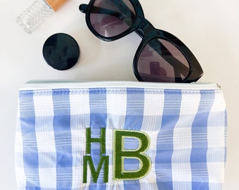 May include: A blue and white gingham makeup bag with the initials "HM" embroidered in green. The bag is lying flat on a white surface with a pair of black sunglasses and a small glass bottle with a wooden top.