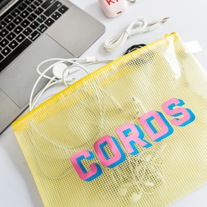May include: A yellow mesh zippered pouch with the word "CORDS" printed in pink and blue letters. The pouch is filled with various white cords and a white Apple Watch charger.