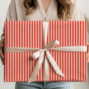 Könnte beinhalten: Ein verpacktes Geschenk mit roten und weißen vertikalen Streifen, gebunden mit einem beigen Band und einer Schleife. Eine Rolle passendes Geschenkpapier, eine Schere und Klebeband liegen auf einer Holzoberfläche.
