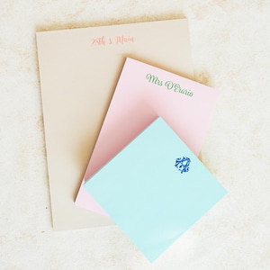 May include: Three personalized notepads with different designs. The top notepad is beige with the text "28th & Main" in pink. The middle notepad is pink with the text "Mrs. D'Eario" in green. The bottom notepad is light blue with a blue monogram.