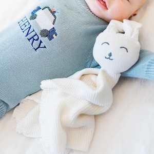 May include: A light blue long-sleeved knit sweater featuring an embroidered police car and the name HENRY in dark blue thread, paired with a white knit security blanket shaped like a rabbit head with embroidered facial features.