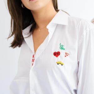 May include: A white button-down shirt featuring embroidered New York City themed patches including a green Statue of Liberty, a red heart with NYC text, a slice of pizza, and a yellow taxi. The letters K, M, and V are embroidered in red vertically.