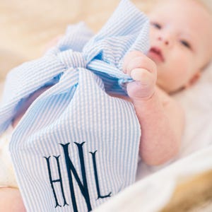 May include: A blue and white striped seersucker baby burp cloth with navy blue embroidered initials. The text "shown as FONT #2 NAVY THREAD" is visible in the image. The burp cloth is tied in a bow.