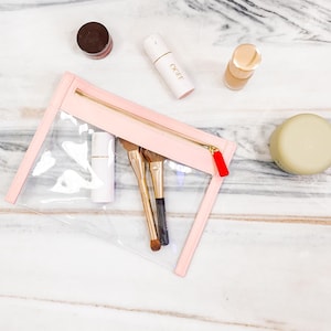 May include: A rectangular clear vinyl pouch with light pink trim and a gold zipper featuring a red pull tab. Inside the pouch are two makeup brushes and a white cylindrical container. Nearby are a small brown jar, a white OGEE tube, and a bottle.
