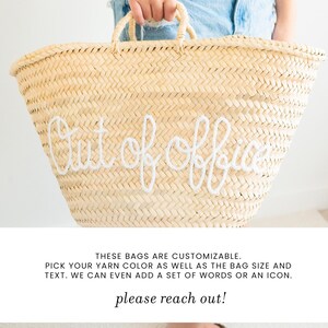 May include: A large, woven straw tote bag with the words "Out of Office" embroidered in white. The bag has two handles and is held by a person wearing denim shorts. The bag is a natural beige color.