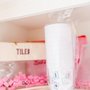 May include: A stack of white plastic cups with a floral design in a clear plastic bag. The cups are on a shelf with a box labeled "TILES" and pink game pieces.