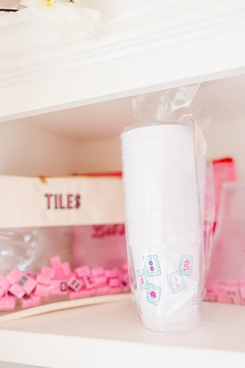 May include: A stack of white plastic cups with a floral design in a clear plastic bag. The cups are on a shelf with a box labeled "TILES" and pink game pieces.