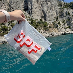 May include: A white mesh bag with the word "SPF" in red, 3D block letters. The bag is held up against a backdrop of a rocky cliffside and blue water. The bag contains rolled-up items.