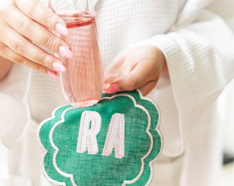 May include: A green linen coaster with a scalloped edge and the letters "RA" embroidered in pink.