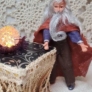 May include: A miniature wizard figurine wearing a pointed hat and a long, white beard, standing in front of a small, ornate table with a crystal ball on top. The table has a fringed fabric skirt and is decorated with a black and silver floral pattern. There are several small, decorative items scattered around the table, including a silver coin, a small knife, and a chain.