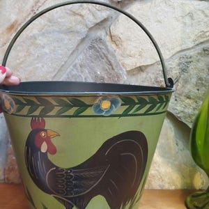 May include: A metal bucket with a black handle and a green exterior. The bucket features a painted rooster design with a red comb and wattle. Floral and leaf patterns decorate the top and bottom of the bucket.