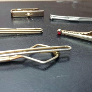 May include: Five vintage tie clips in gold and silver tones are arranged on a dark surface. The clips vary in design, including a gold clip with a red gemstone and a silver clip with a simple bar design. Accessories.
