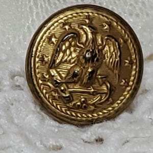 May include: A gold-colored metal button with an eagle design and a rope-like border. The eagle is holding a branch in its talons and there are stars around the eagle.